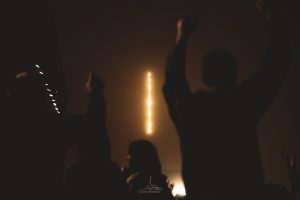 Xente levantando as mans coa chimenea da fábrica azucreira de Portas co letreiro luminos de Portamérica de fondo durante o festival Portamérica 2026